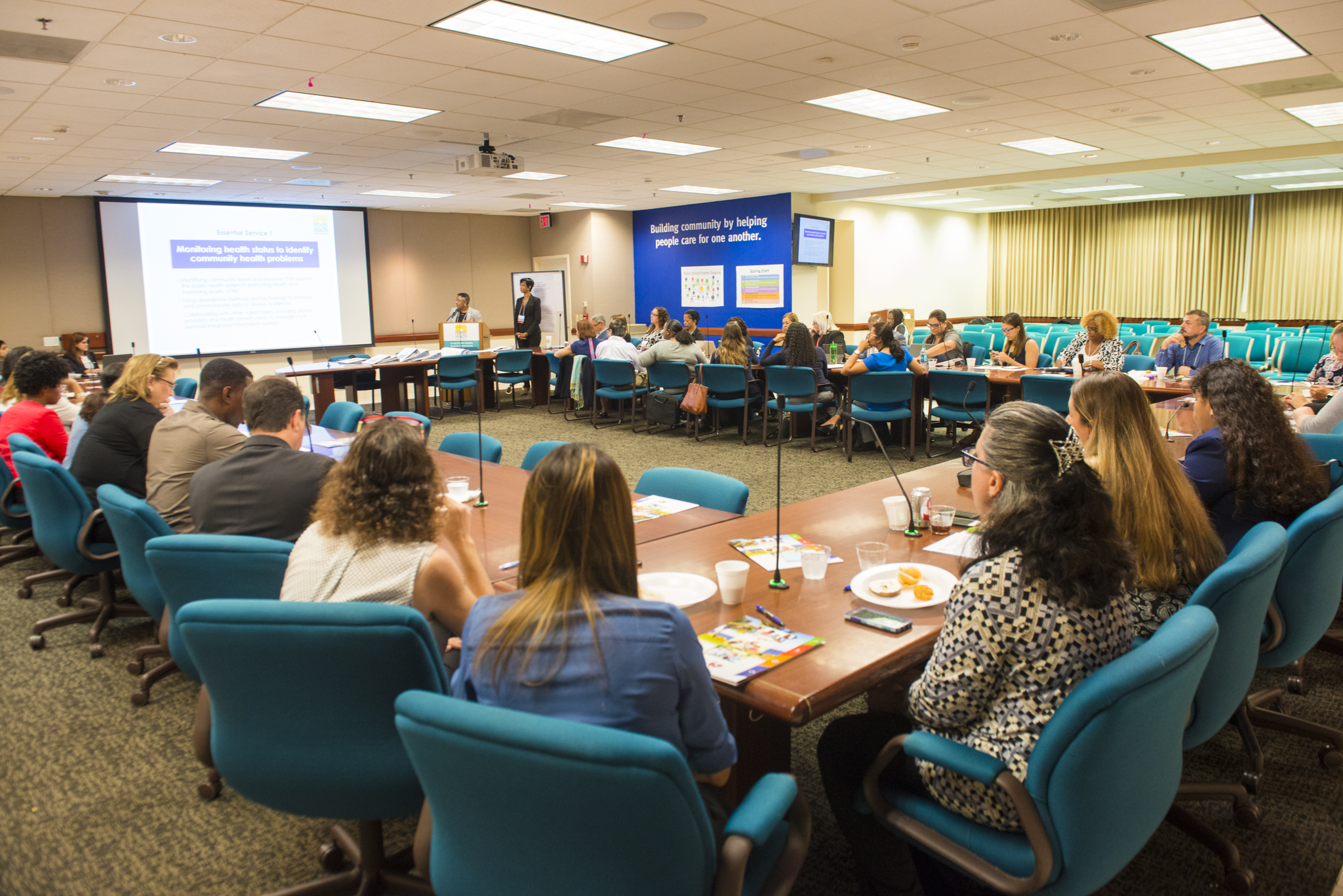 A photo of a conference room in which people sit at a table in a square formation in front of a projector screen